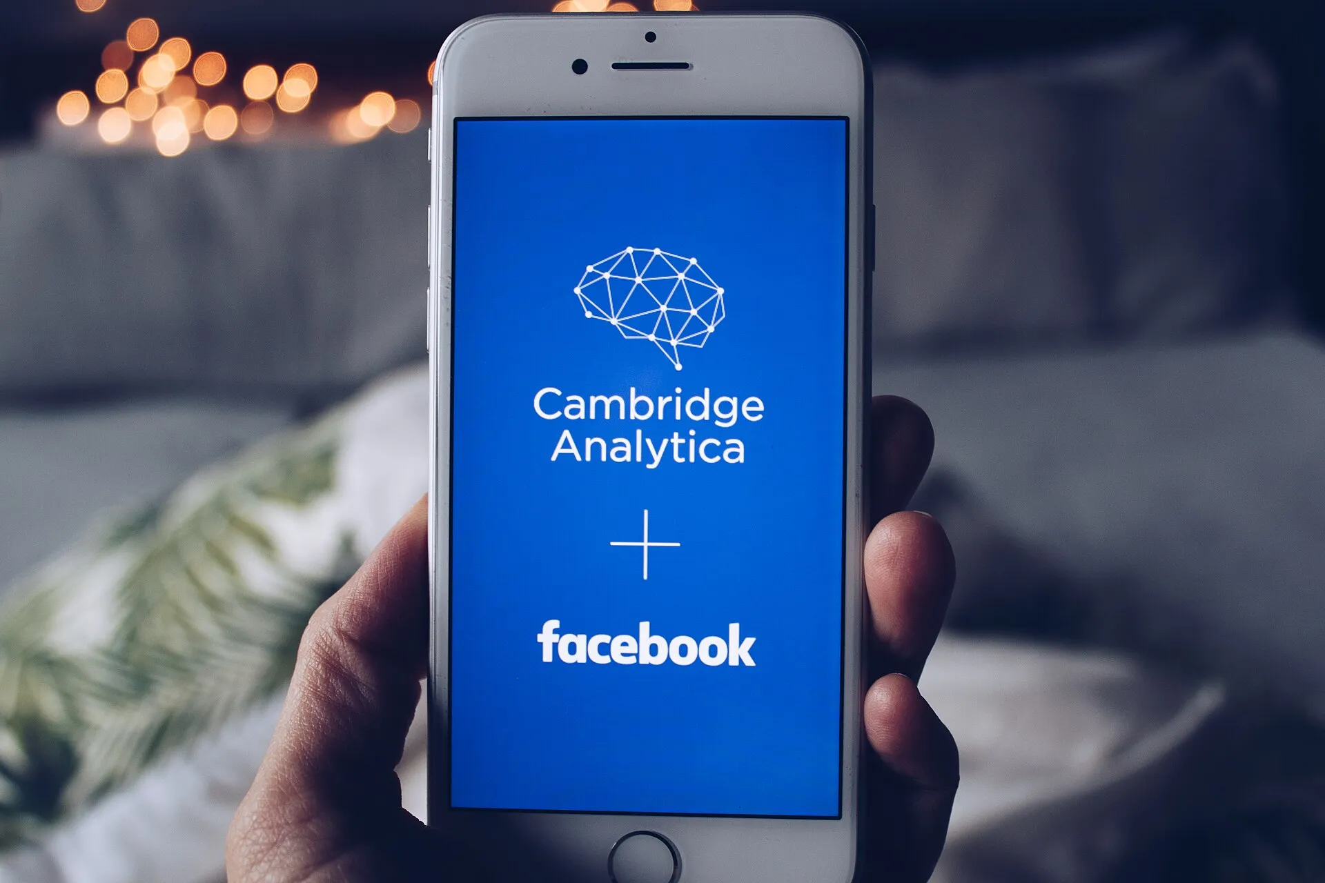Mão a segurar um telemóvel cujo ecrã azul mostra o logótipo da Cambridge Analytica, um sinal de mais e o logótipo do Facebook.